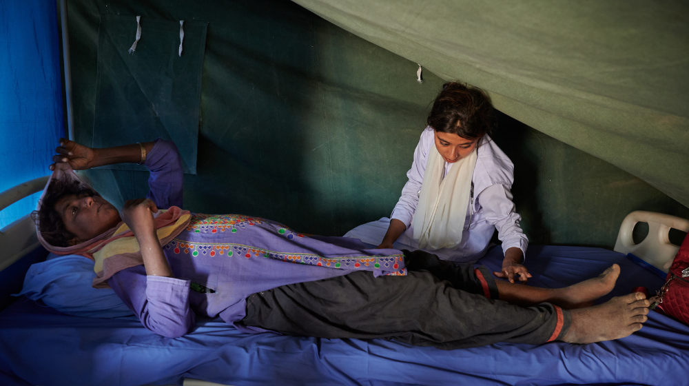 Une femme enceinte portant une tunique violette est allongée sur un lit d’hôpital dans une tente verte.  Une femme assise sur le lit d’à côté examine ses jambes à la recherche de signes d’œdème.