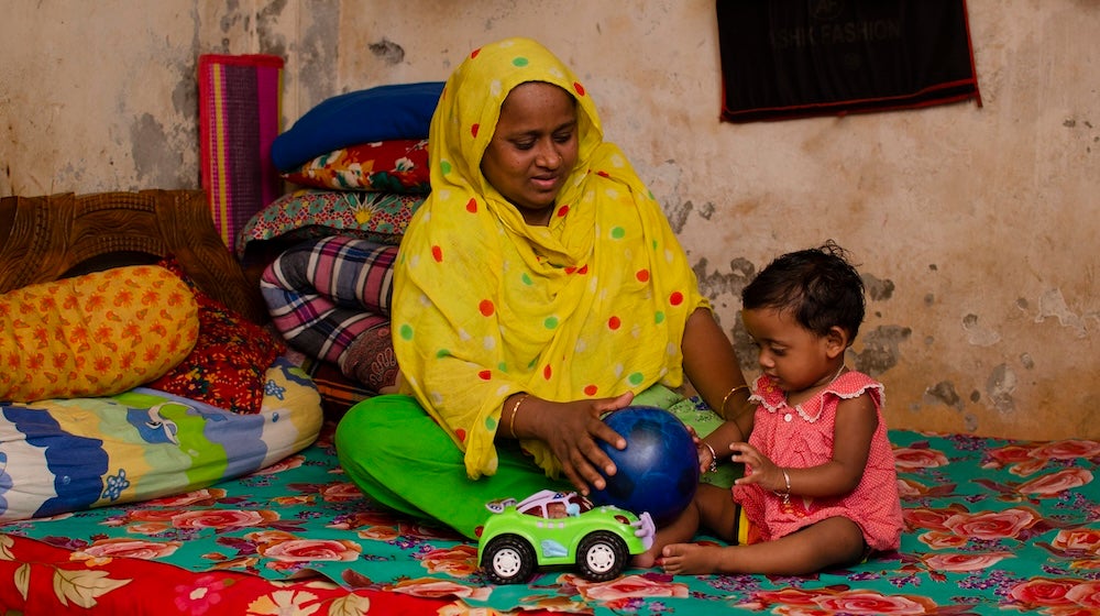 A mother plays with her child while sitting on a bed.
