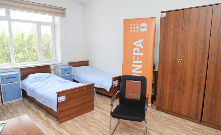 Una habitación con dos camas individuales, mesitas de\ noche, un armario, una silla y una bandera del UNFPA. Detrás de las camas, una ventana con vista a unos árboles.