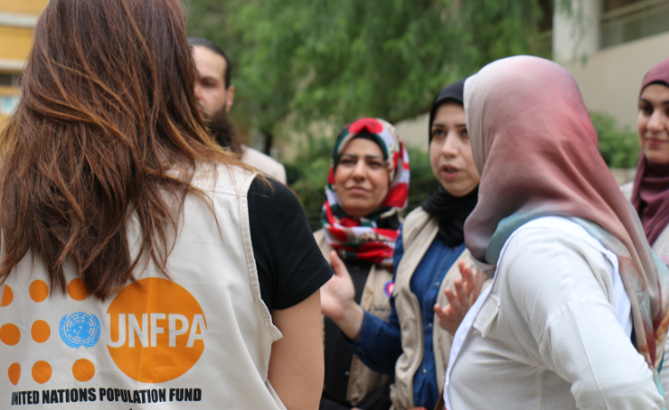 Una mujer con un chaleco con el logotipo del UNFPA habla con un grupo de mujeres y un hombre en un patio al aire libre.