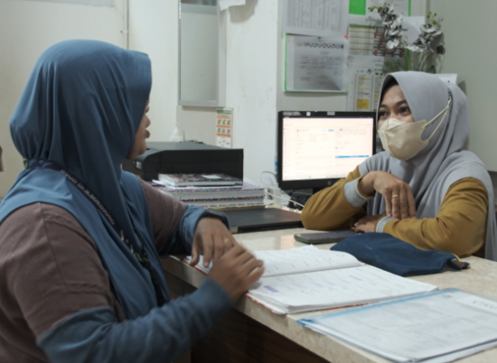 Two women in head coverings sit across from one another in a medical office with a computer in the background and files open in front of them