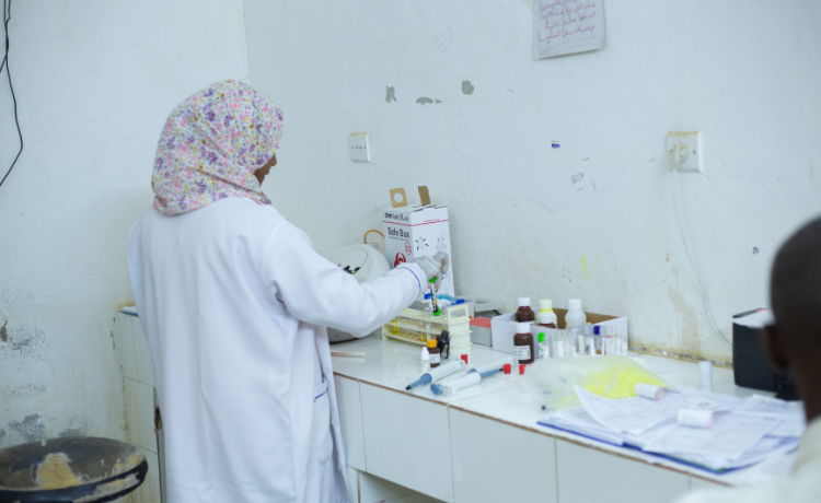  عاملة صحية ترتدي معطفا أبيض وغطاء رأس منمق تحمل عينة دم أمام مواجهة الحائط داخل عيادة