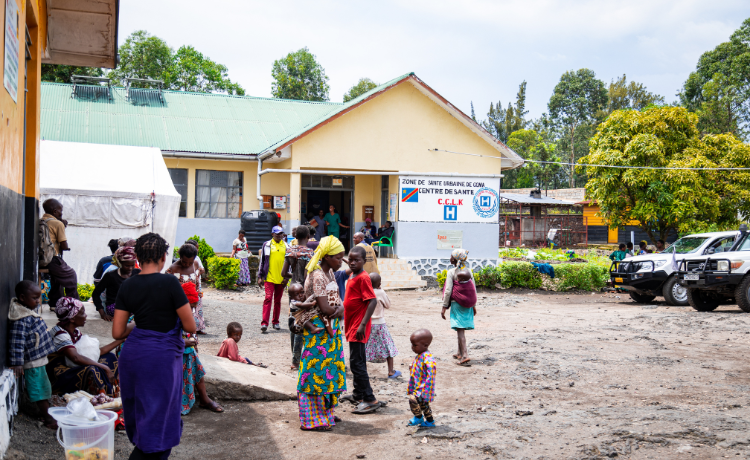 Photo d’une clinique médicale devant laquelle sont garées deux voitures. Au premier plan, plusieurs personnes attendent.