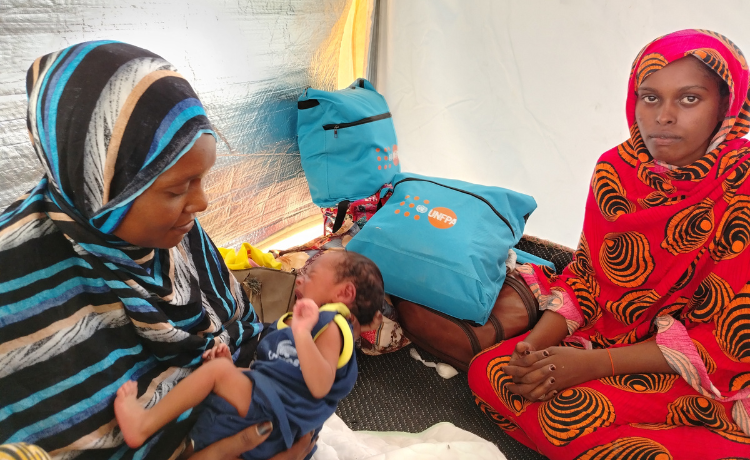  Une femme portant un voile à rayures tient un nouveau-né ; elle est assise sur le sol dans une tente, à côté d’une autre femme et de kits de dignité au logo de l’UNFPA.
