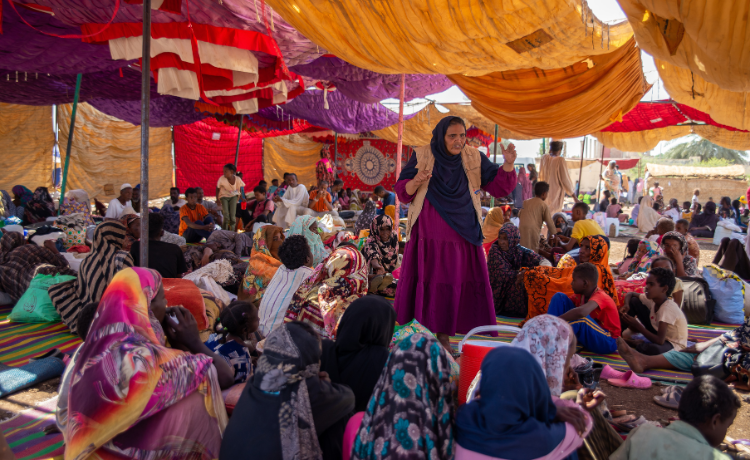 Una mujer con un chaleco del UNFPA se dirige a una multitud de mujeres sentadas debajo de una tienda de campaña