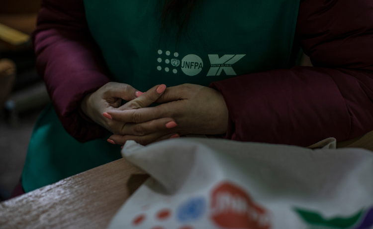 Une femme portant du vernis à ongles orange croise les mains sur ses genoux ; elle est assise à un bureau, et porte un haut vert sur lequel on distingue le logo de l’UNFPA
