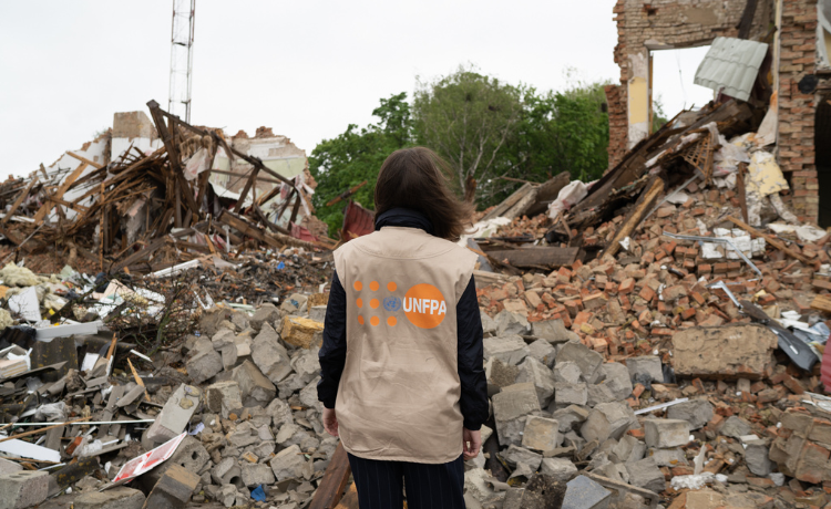 Une femme se tient devant des maisons en ruines dont il ne reste que des gravats ; elle est dos à l’objectif et porte un gilet de l’UNFPA
