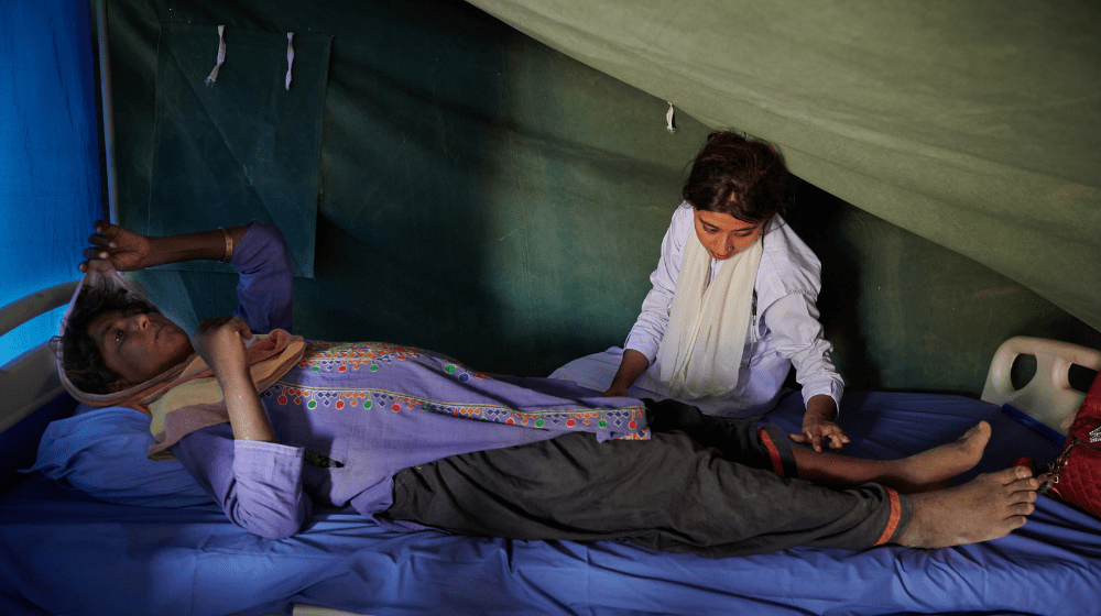 Une femme enceinte portant une tunique violette est allongée sur un lit d’hôpital dans une tente verte.  Une femme assise sur le lit d’à côté examine ses jambes à la recherche de signes d’œdème.