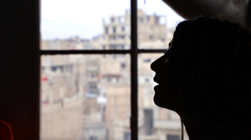 Una mujer retroiluminada de perfil junto a una ventana, a través de la cual se aprecian edificios destruidos