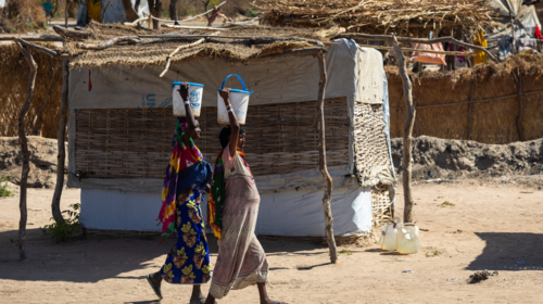 Dos mujeres pasan frente a un refugio improvisado llevando cubos del ACNUR en la cabeza
