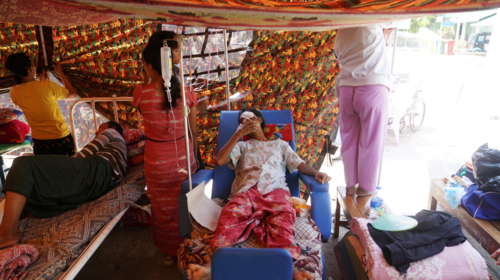 Una mujer en una cama de hospital dentro de una tienda improvisada, al lado de un hombre también en cama y tres mujeres colgando material para darles sombra