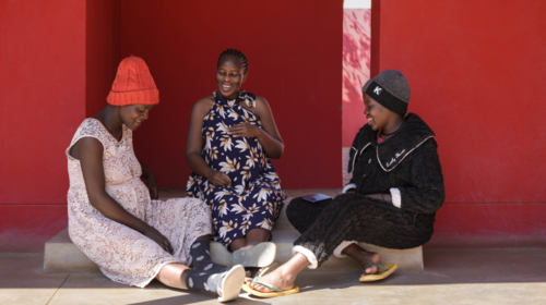 Three pregnant women sit on a step outside against a red wall laughing